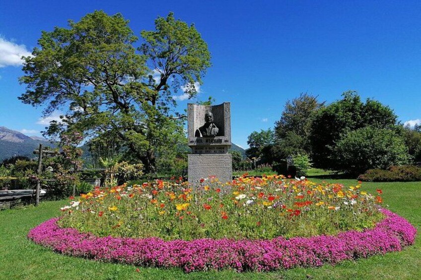 A statue surrounded by a circular flower bed with various colors, set in a park with trees and mountains in the background. 1