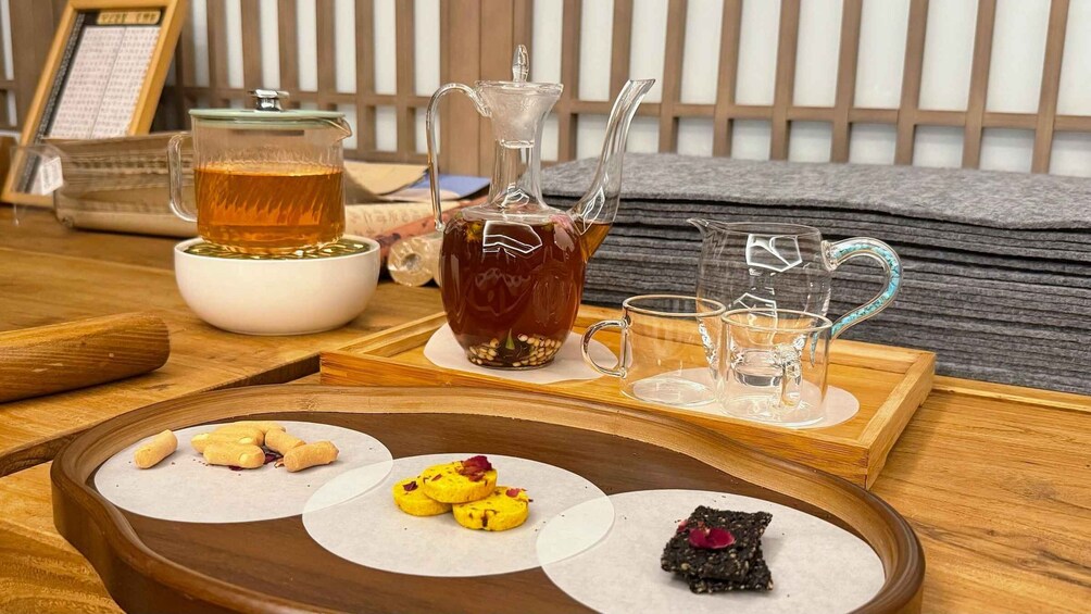 A tea set with a glass pitcher, cups, and a teapot on a wooden tray with snacks. 3