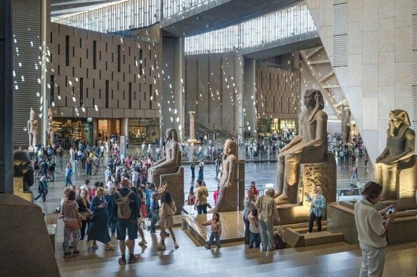 A spacious indoor area with large golden statues of Egyptian figures, modern architecture, and a crowd of visitors. 1