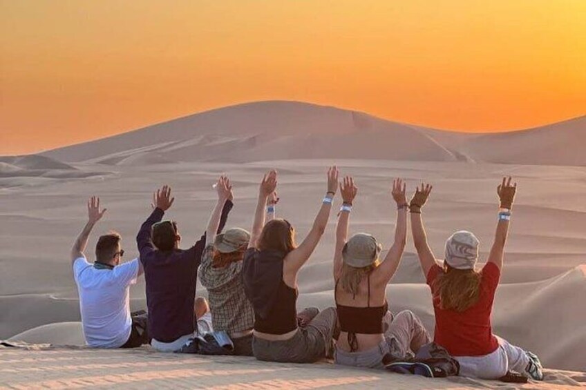 Eine Gruppe von Menschen sitzt auf Sanddünen, die Hände zum Himmel erhoben, und blickt auf den Sonnenuntergang. 2