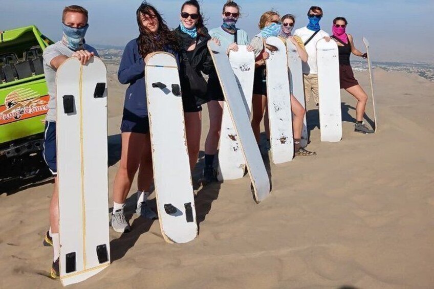 Eine Gruppe von Menschen trägt Surfbretter an einem sandigen Strand, im Hintergrund ist ein grünes Fahrzeug zu sehen. 1