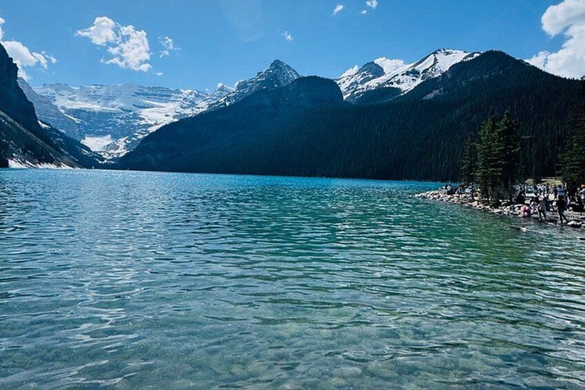 A clear lake surrounded by mountains with a rocky shoreline and a few people. 4