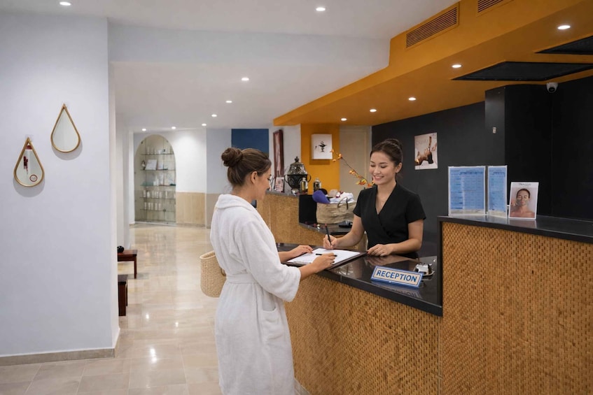 A reception desk with a sign, a woman in a robe, and another woman behind the counter. 1