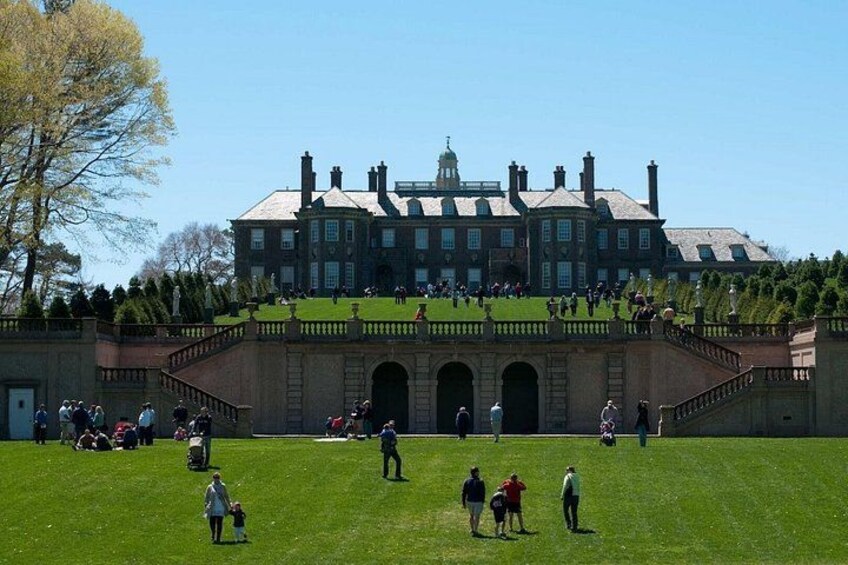 Un gran edificio histórico con un césped verde frente a él, personas caminando y sentadas sobre el césped, y un puente de piedra que conduce al edificio. 1
