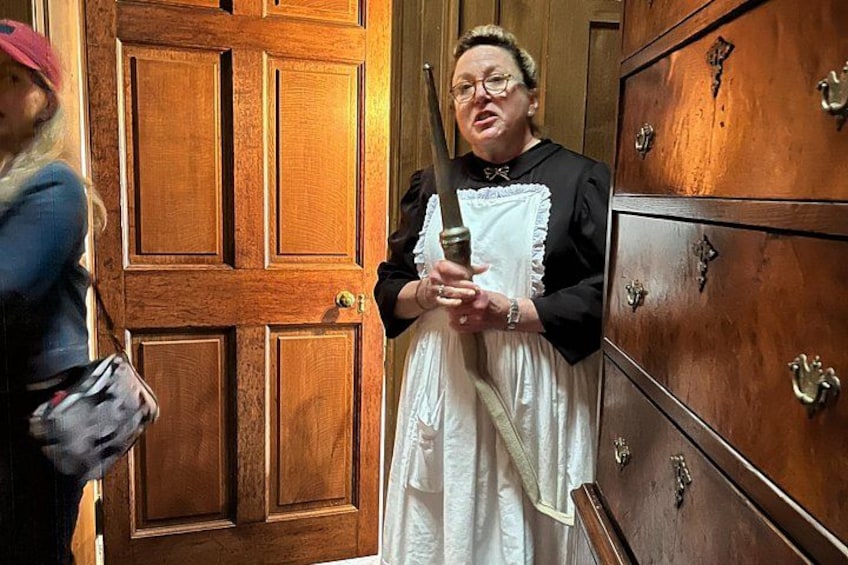A person in a white dress holding a large knife, standing in front of a wooden door with brass handles. 5