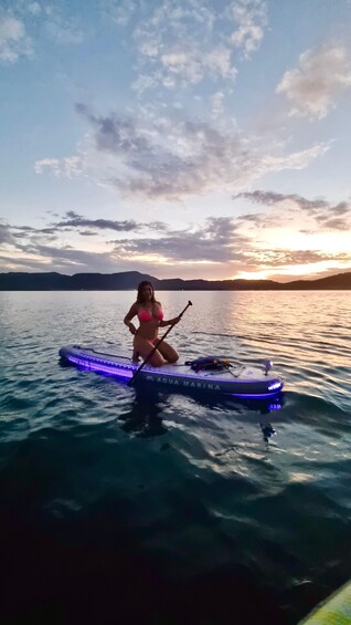 A person in a pink bikini paddling a kayak with blue lights on a calm body of water at sunset. 5