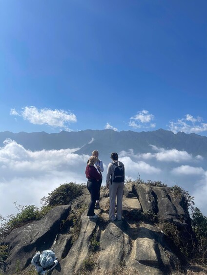 Two people standing on a rocky outcrop overlooking a mountain range and clouds below. 5