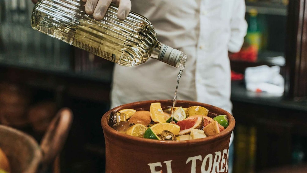 A person pouring a clear liquid into a ceramic pot filled with various fruits and spices. 4