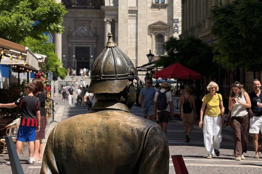 Une statue de bronze représentant un homme portant un casque, une rue animée où l’on voit des piétons, et un bâtiment à l’architecture classique en arrière-plan. 5