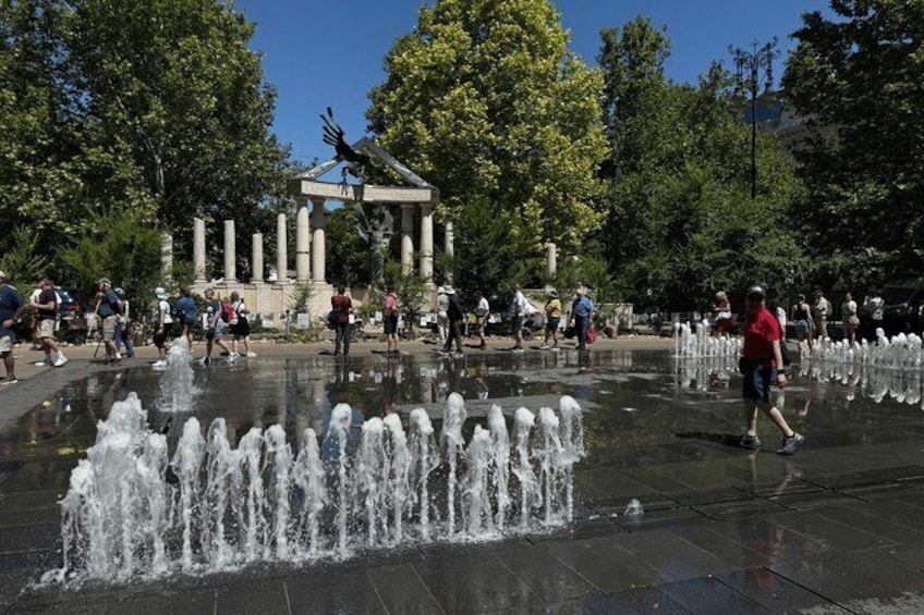 A public fountain with water jets, surrounded by a plaza with people walking and a classical monument in the background. 3