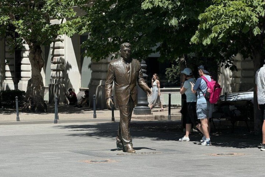 A statue of a man in a suit on a city street with trees and people around. 4