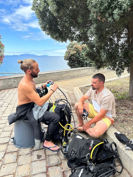 Two men sitting on a stone bench by the sea, with scuba diving gear and a backpack on the ground. 2
