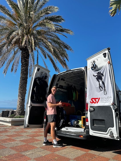A person standing next to a van with open doors, displaying scuba diving equipment and a banner for Scuba Schools International. 3