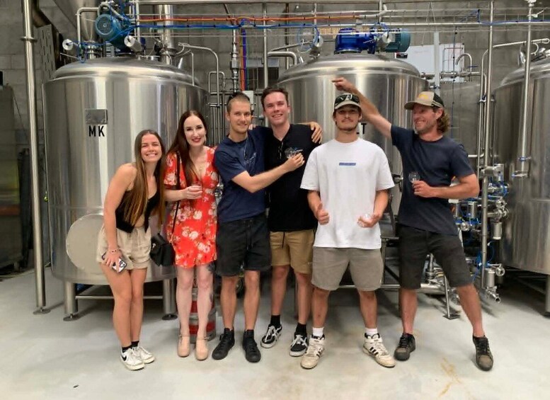 A group of people standing in a brewery with stainless steel tanks and pipes in the background. 1