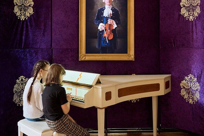 Två personer spelar på ett stränginstrument vid ett piano, och ovanför dem hänger ett porträtt inramat. 2