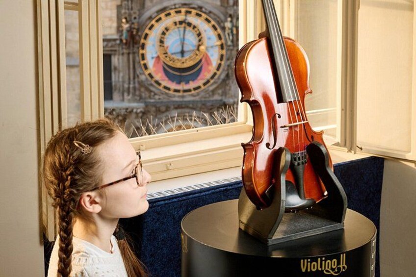 En fiol står på ett notställ, och framför ett fönster med utsikt över en klocka och en byggnad finns en dekorativ platta med texten ”Violino”. 1