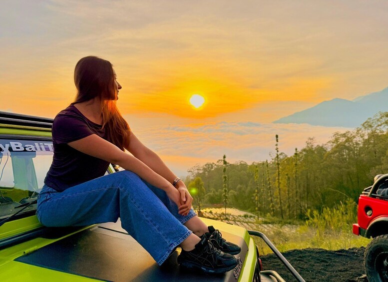 A person sitting on a vehicle's roof, overlooking a sunset with mountains in the distance. 1