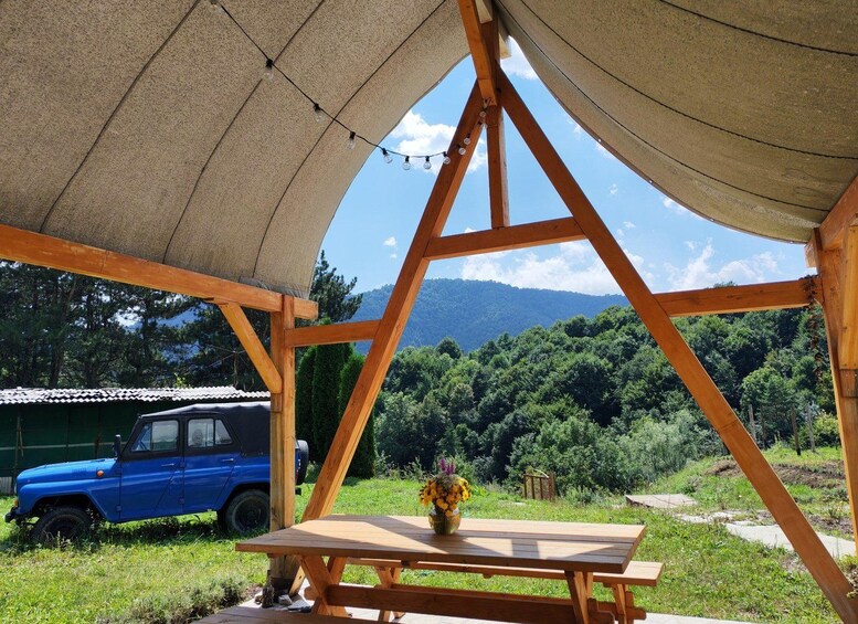 A wooden pavilion with a table and a vase of flowers, a blue car, and a mountainous landscape in the background. 2
