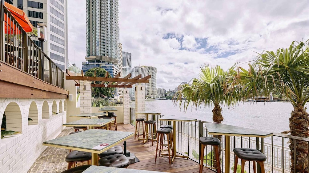 An outdoor bar with tables and stools overlooking a body of water, with tall buildings in the background. 3