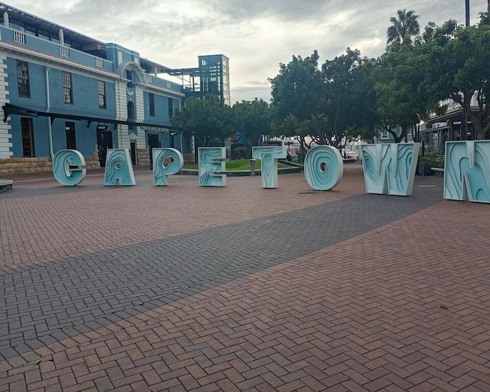 Een geplaveid plein met grote, lichtblauwe letters die 'CAPETOWN' spellen en een historisch gebouw op de achtergrond. 1