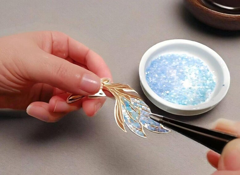 A hand holding a gold leaf-shaped pendant with blue accents, being cleaned with a brush and a bowl of blue powder. 2
