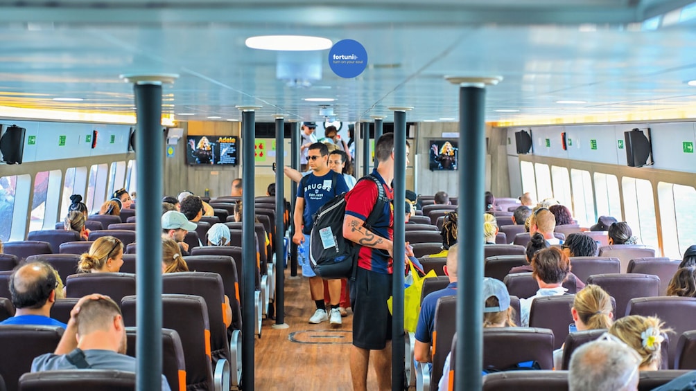 A ferry interior with passengers seated, a man in a red and blue shirt standing, and a blue sign overhead. 3