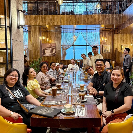 A group of people seated around a long wooden table in a restaurant with hanging lights and a large window in the background. 3