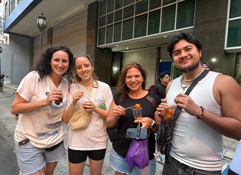 Four people standing on a sidewalk, holding drinks and snacks, with a building in the background. 5
