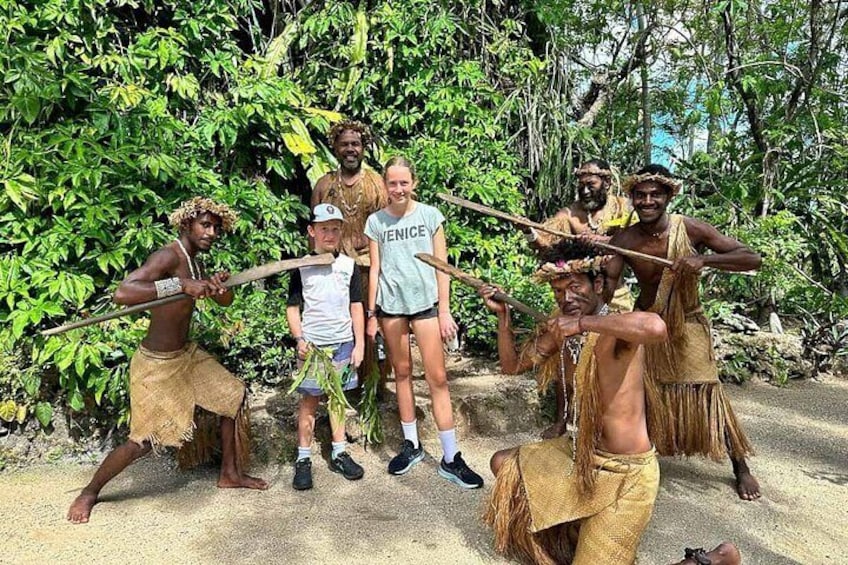 A group of people in traditional attire, some holding spears, standing in a natural, forested area. 1