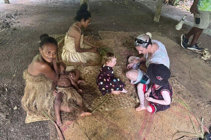 A group of people sitting on mats outdoors, some children and adults, in a casual, communal setting. 2