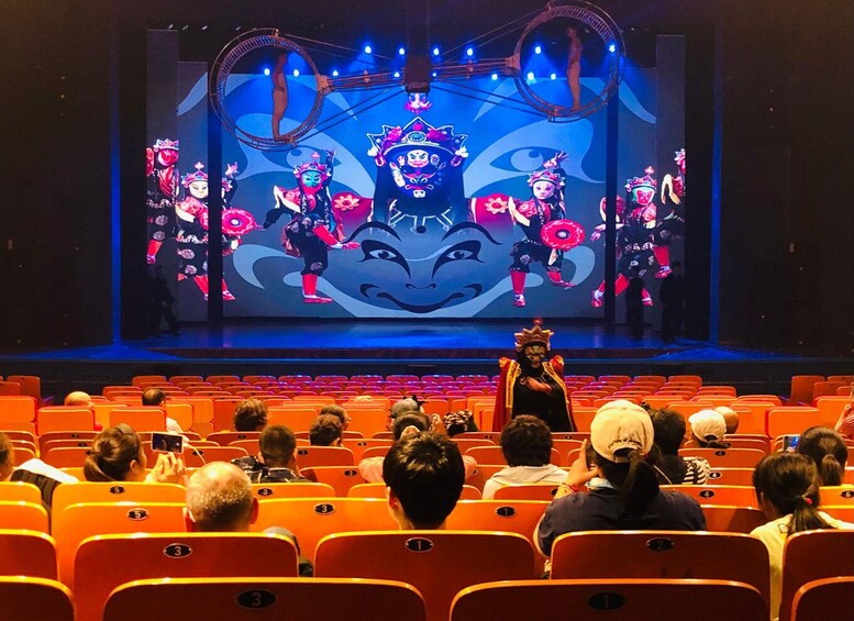 A theater with a stage performance, featuring performers in traditional costumes and a large screen displaying a cultural scene. 1
