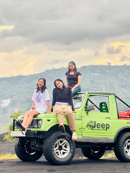 Three people sitting on a green Jeep with 'Jeep Adventure' written on it, parked on a road with a hilly landscape in the background. 5