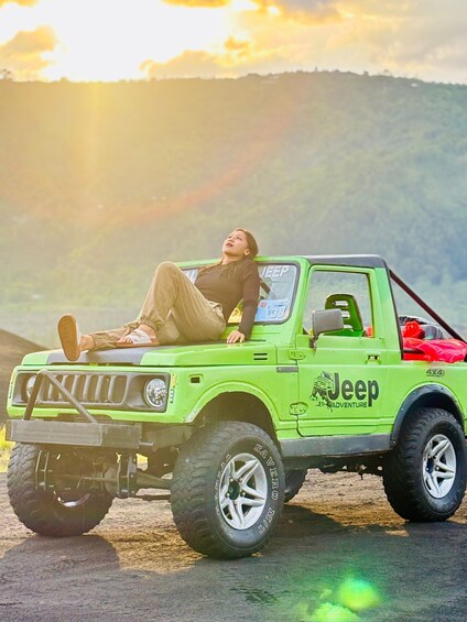 A person reclining in a green Jeep 4x4 with 'Jeep Adventure' branding, parked on a dirt road with a mountainous backdrop. 4