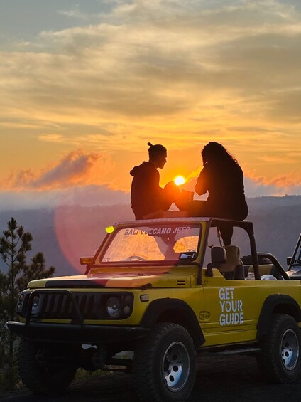 A yellow jeep with 'BALI VOLCANO JEEP' and 'GET YOUR GUIDE' on the side parked on a mountain overlooking a sunset. 1