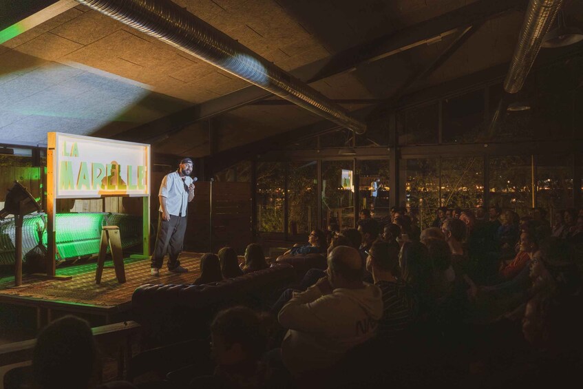 A person speaking to an audience in a dimly lit room with a stage and a large screen displaying 'LA MARIE.' 3
