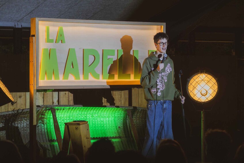 A person standing on a stage with a microphone, a sign reading 'LA MARELLA,' and a green-lit structure behind them. 1