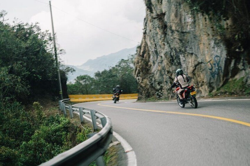 Una strada tortuosa con un motociclista e un altro motociclista più avanti, immersa nel verde e con una parete rocciosa. 2