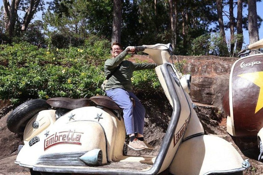 A person on a vintage vehicle with a logo, surrounded by greenery and trees. 3