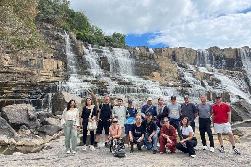 A group of people posing in front of a waterfall with rocky terrain and greenery in the background. 4