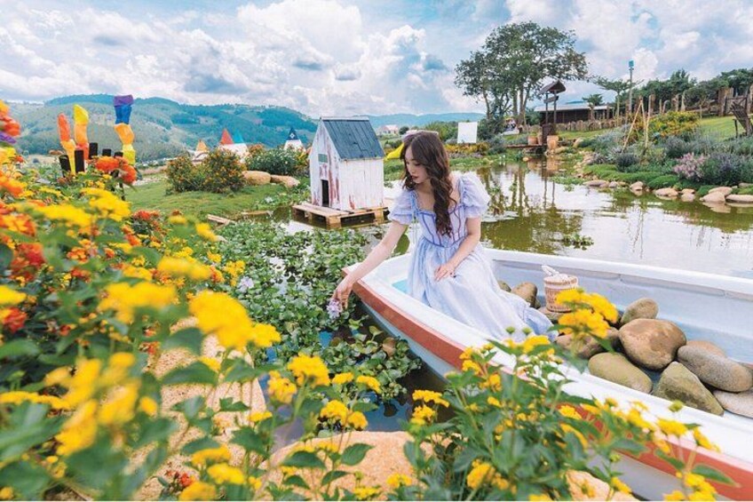 A woman in a white dress by a pond with flowers, a small house, and a rural landscape. 2
