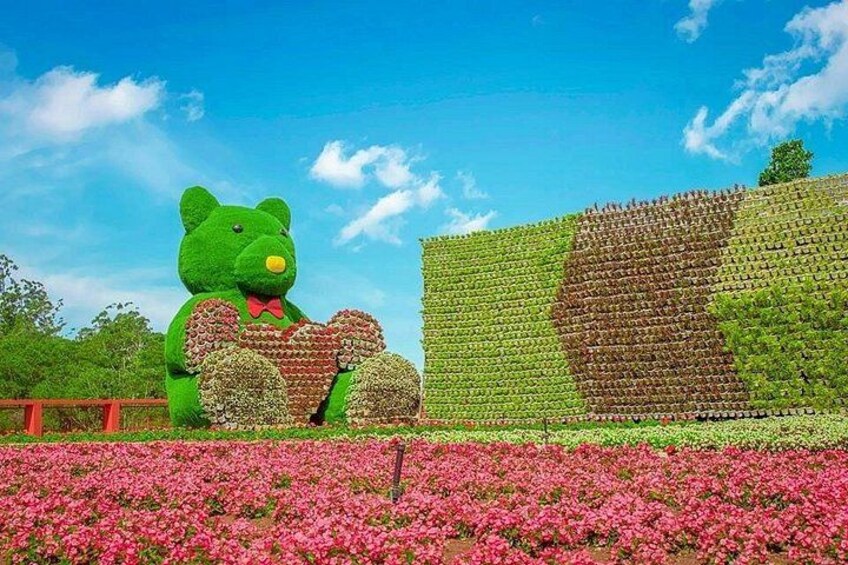 A large green teddy bear sculpture, a wall of green and brown plants, and a field of pink flowers. 4