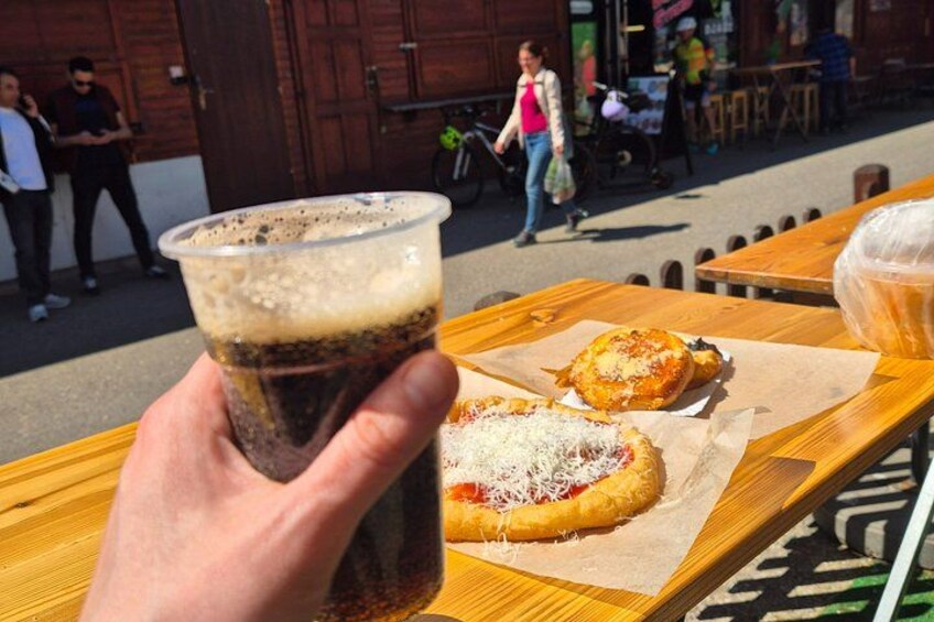 Een hand die een bekertje soda vasthoudt, een pizza op een houten tafel en een persoon op de achtergrond. 4