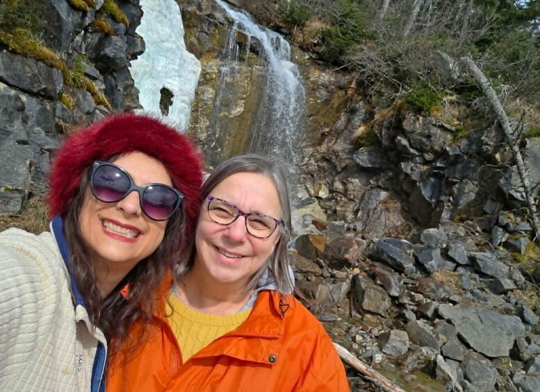 Two people in front of a waterfall with rocky terrain and sparse vegetation. 4