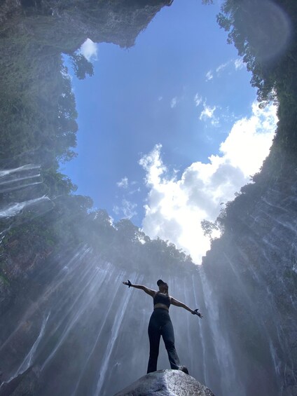 A person standing on a rock with arms outstretched, looking up at a waterfall and the sky. 1