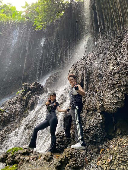 Two people posing in front of a waterfall with lush greenery in the background. 3