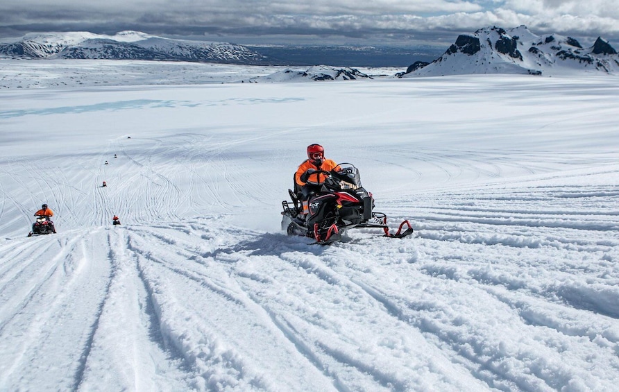A snowmobile on a snowy landscape with tracks, a person in orange, and distant mountains. 3