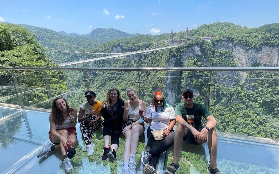 A group of people sitting on a glass floor with a scenic view of a mountainous landscape and a bridge in the background. 5