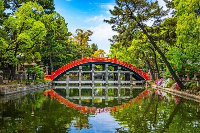 Een rode brug over kalm water, omgeven door weelderige groene bomen en een heldere blauwe lucht. 4