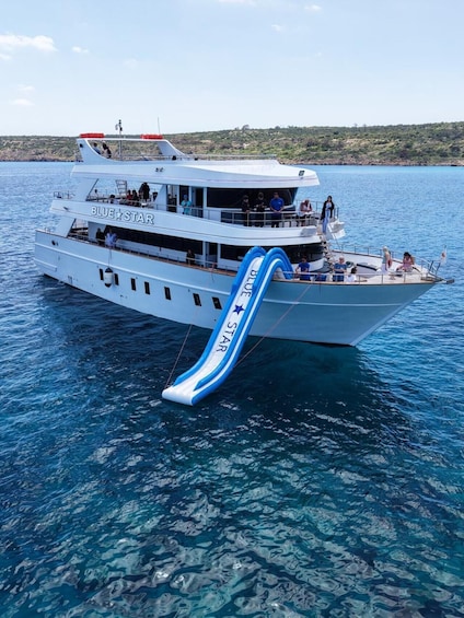 A large white boat with a blue and white slide on the deck, named 'BLUE STAR,' floating on clear blue water. 4