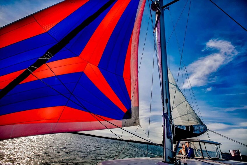 A sailboat with a red and blue sail, a person in a blue jacket, and a flag in the background. 1